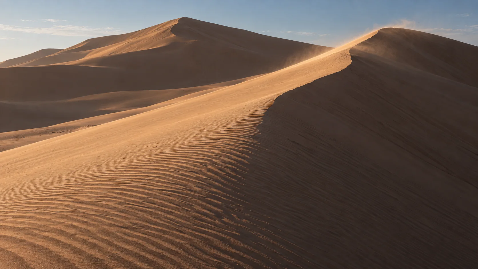Wind carrying sand over the sharp crest of a sunlit dune